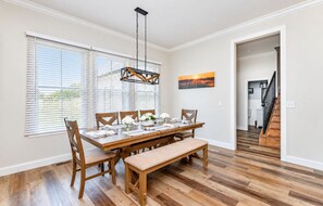 A dining area with a wooden table, chairs, and a bench. There are white walls, a painting, and a staircase in the background.