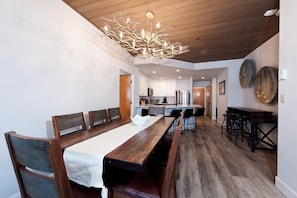 Uma sala de jantar com uma longa mesa de madeira, cadeiras, um candelabro e uma cozinha ao fundo.