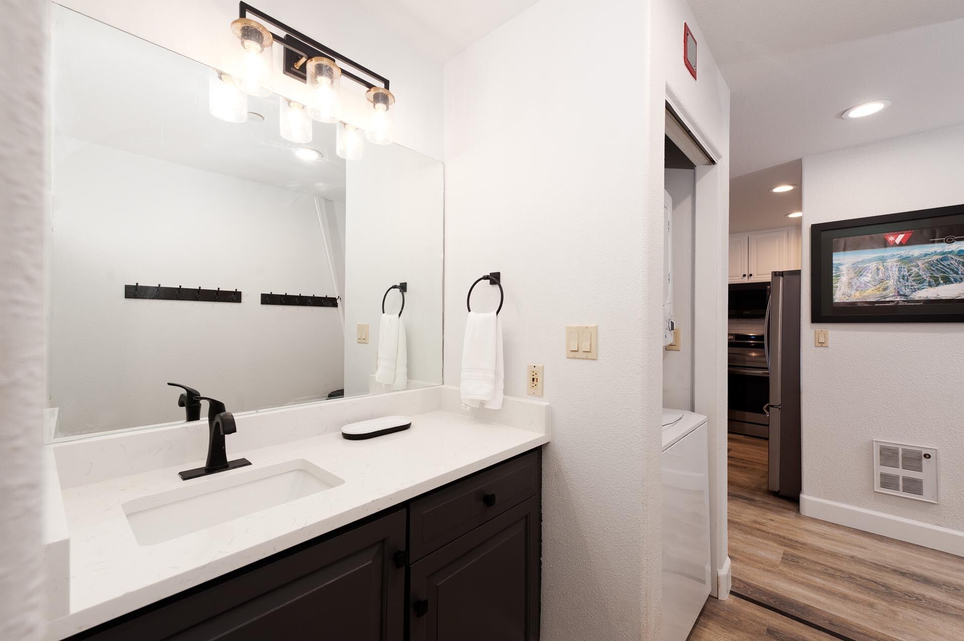 Une salle de bain moderne avec un grand miroir, deux lavabos et un meuble-lavabo. On y trouve également un porte-serviettes et une porte qui donne sur la cuisine.