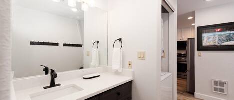 A modern bathroom with a large mirror, two sinks, and a vanity. There is a towel rack and a door leading to a kitchen area.