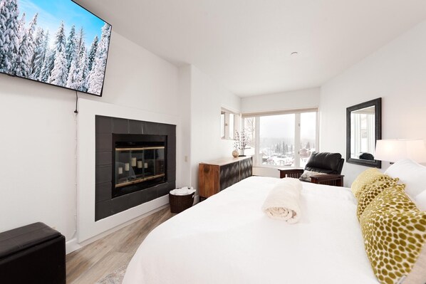 Quarto de hotel moderno com uma cama grande, lareira, vista para uma paisagem nevada e um horizonte urbano pela janela.