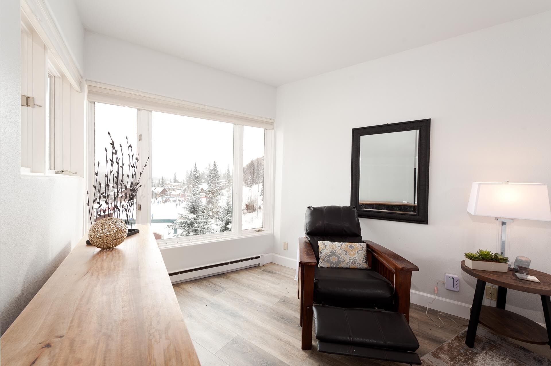 Ein Zimmer mit einer hölzernen Bank, einem schwarzen Ledersessel mit gemustertem Kissen, einem Spiegel an der Wand und einem Fenster mit Blick auf schneebedeckte Bäume.