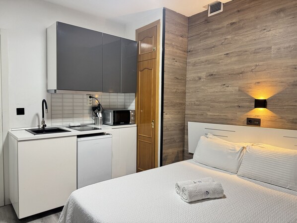 A compact hotel room with a kitchenette, a bed with a neatly folded towel, and a wall-mounted light fixture. - Cosy Room with Bathroom Gran Vía | 034 (Madrid)
