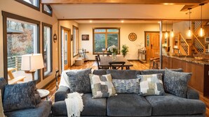A cozy living room with a grey sofa, a dining table, and a kitchen area with a wooden staircase. - Luxury Estate & Rustic Mountain Retreat Asheville (Buncombe County)