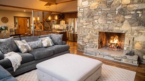 A modern living room with a stone fireplace, a grey sofa, and a kitchen in the background. - Luxury Estate & Rustic Mountain Retreat Asheville (Buncombe County)