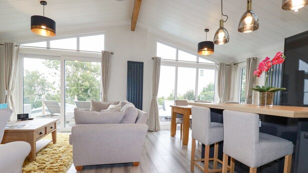 A modern living room with a sofa, dining table, and chairs. There are large windows with curtains, a wooden ceiling, and pendant lights. - Mordros Lodge (Redruth)