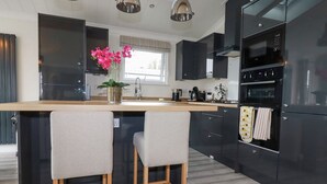 A modern kitchen with a central island, two bar stools, black cabinetry, and a stainless steel range hood. - Mordros Lodge (Redruth)