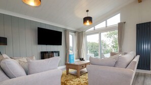 A modern living room with two sofas, a coffee table, a flat-screen TV, and large windows with curtains. - Mordros Lodge (Redruth)