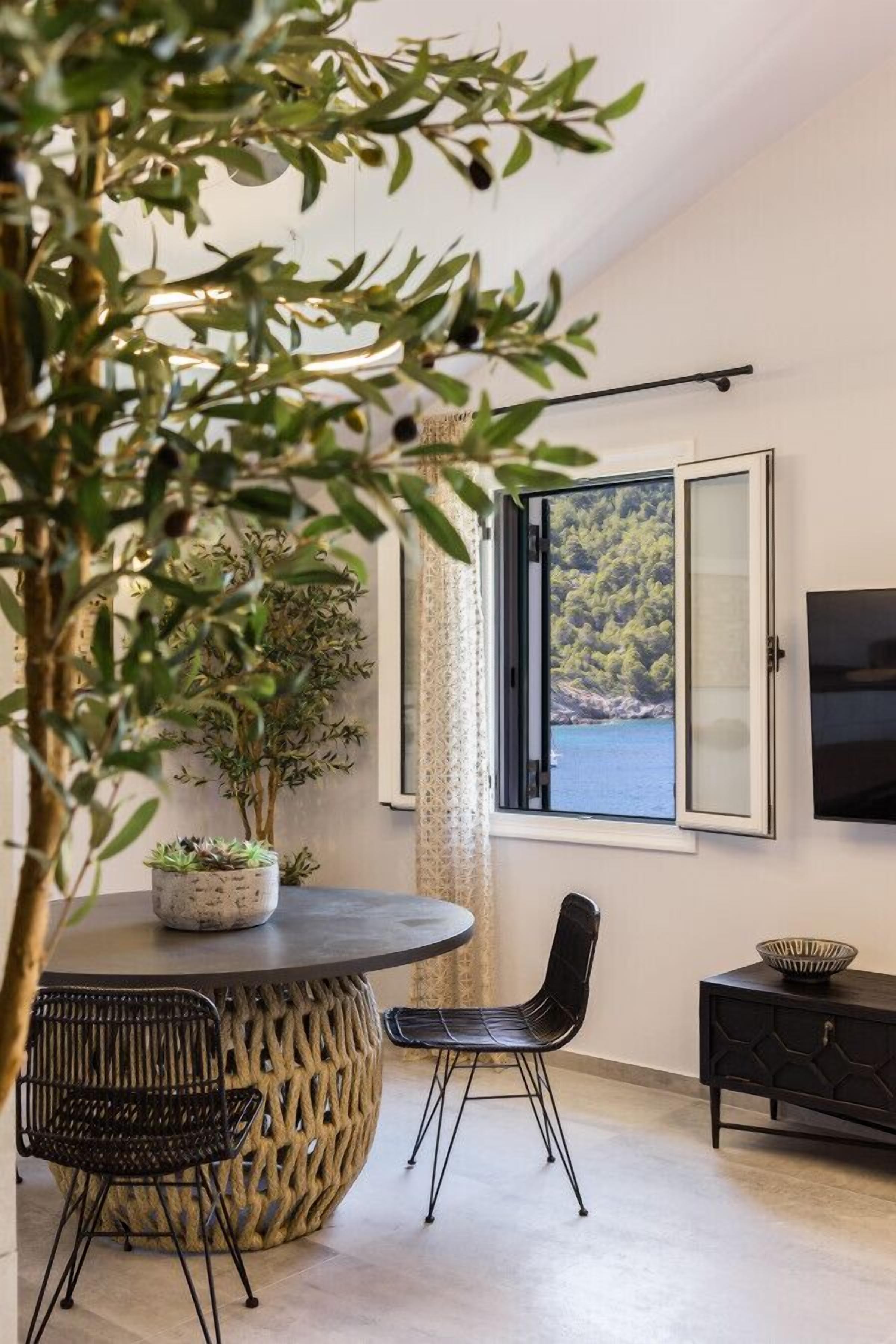 Uma sala de jantar com uma mesa redonda, duas cadeiras, uma planta em vaso e vista para a paisagem costeira através de uma janela.