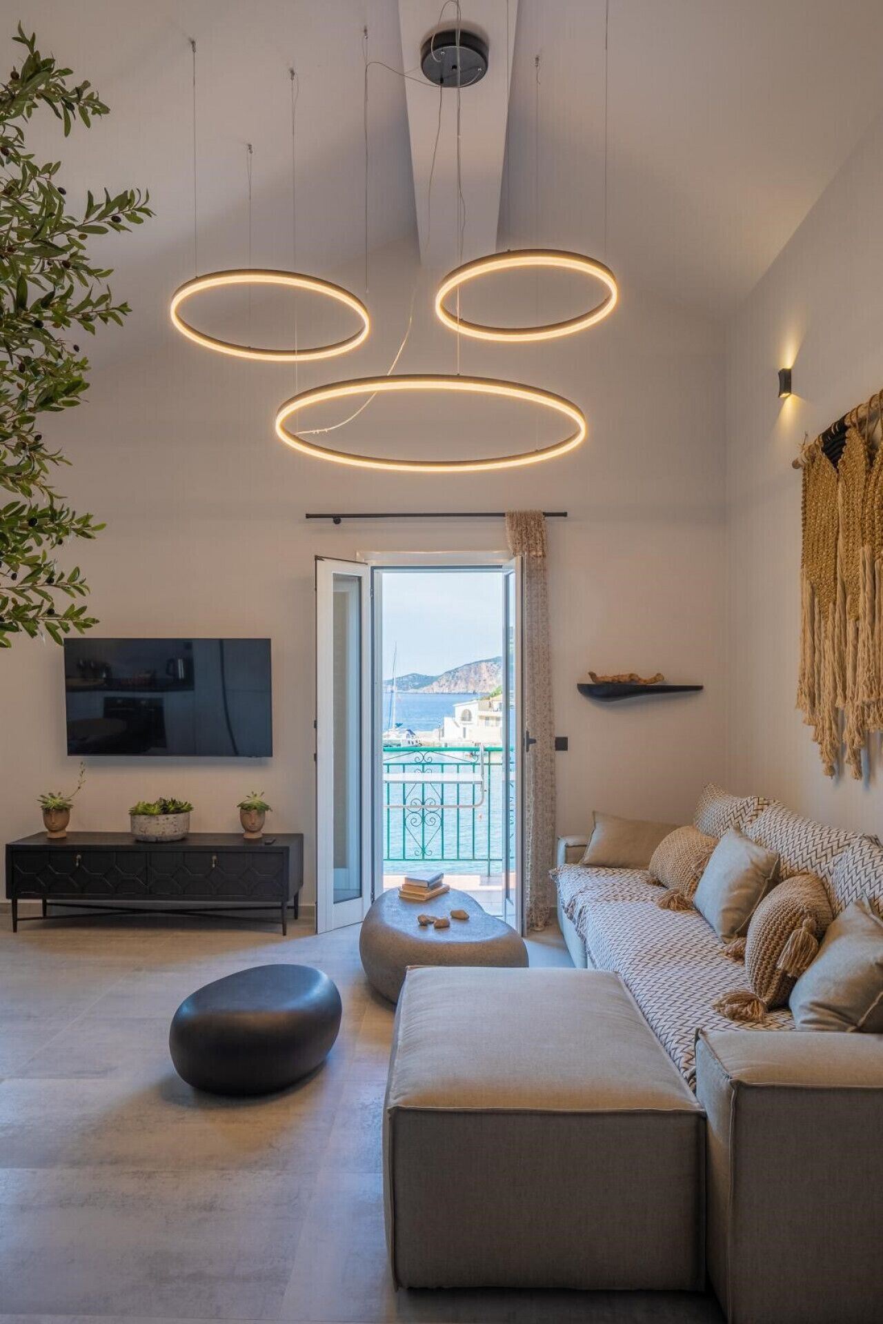 Een moderne woonkamer met een grote televisie, een bank, poefs en uitzicht op zee vanaf het balkon.