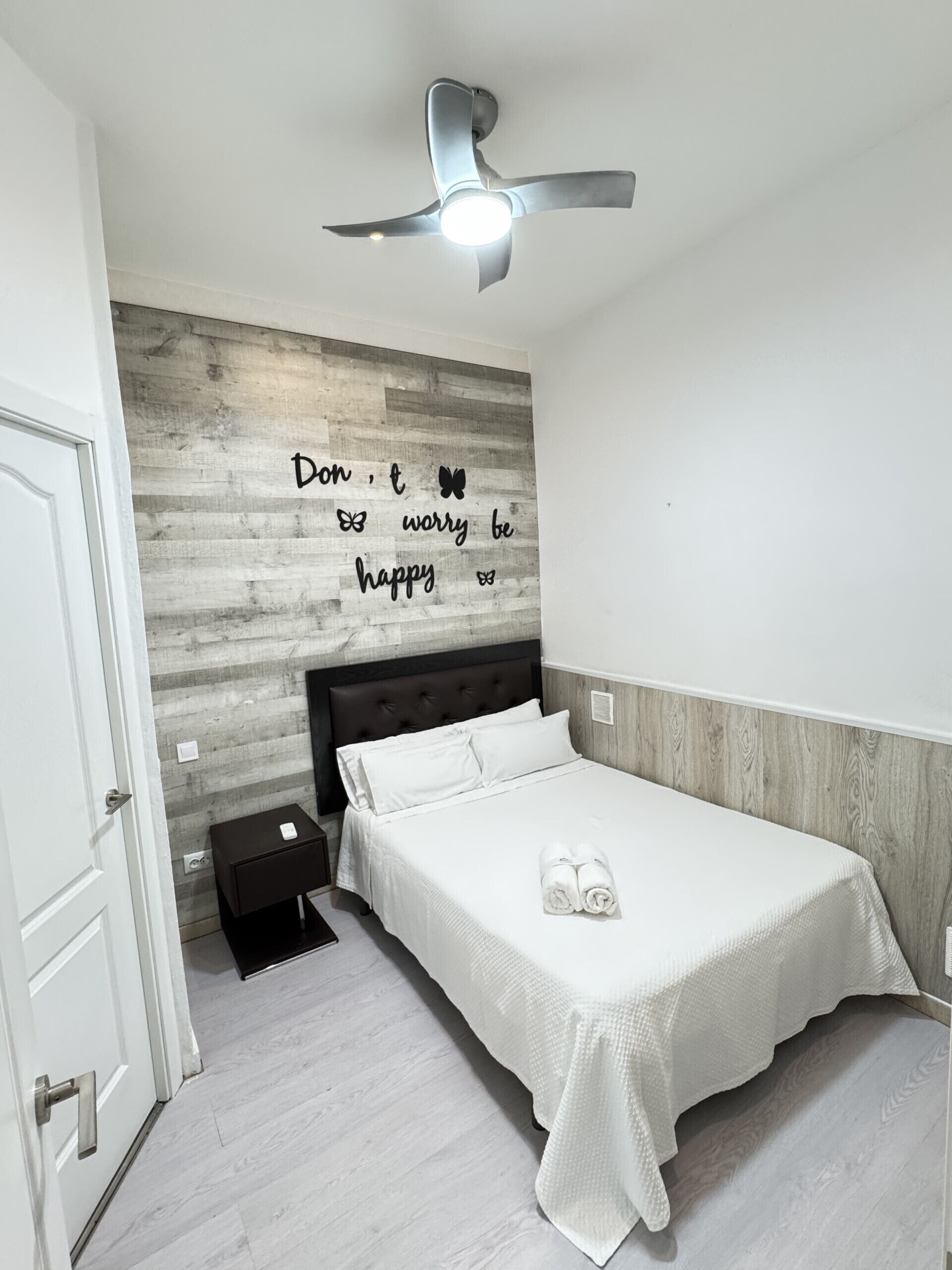 Um quarto com cama, ventilador de teto, parede com frase motivacional e uma pequena mesa de cabeceira.