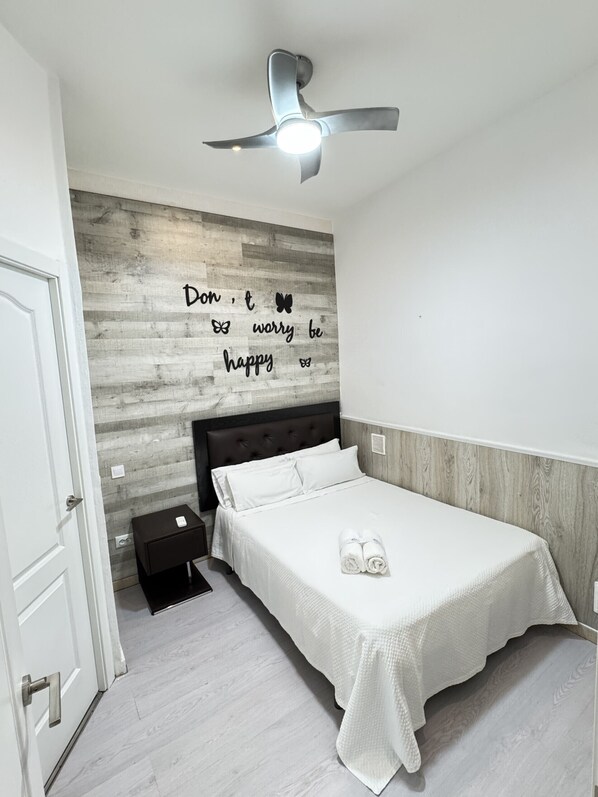 Un dormitorio con cama, ventilador de techo, pared con una cita motivacional y una mesita de noche pequeña.