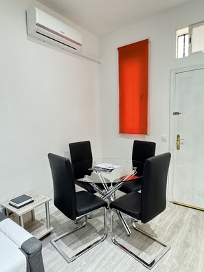 Una habitación pequeña y moderna con una mesa de vidrio y cuatro sillas negras, un sofá blanco, una mesita auxiliar y una pared de acento roja.