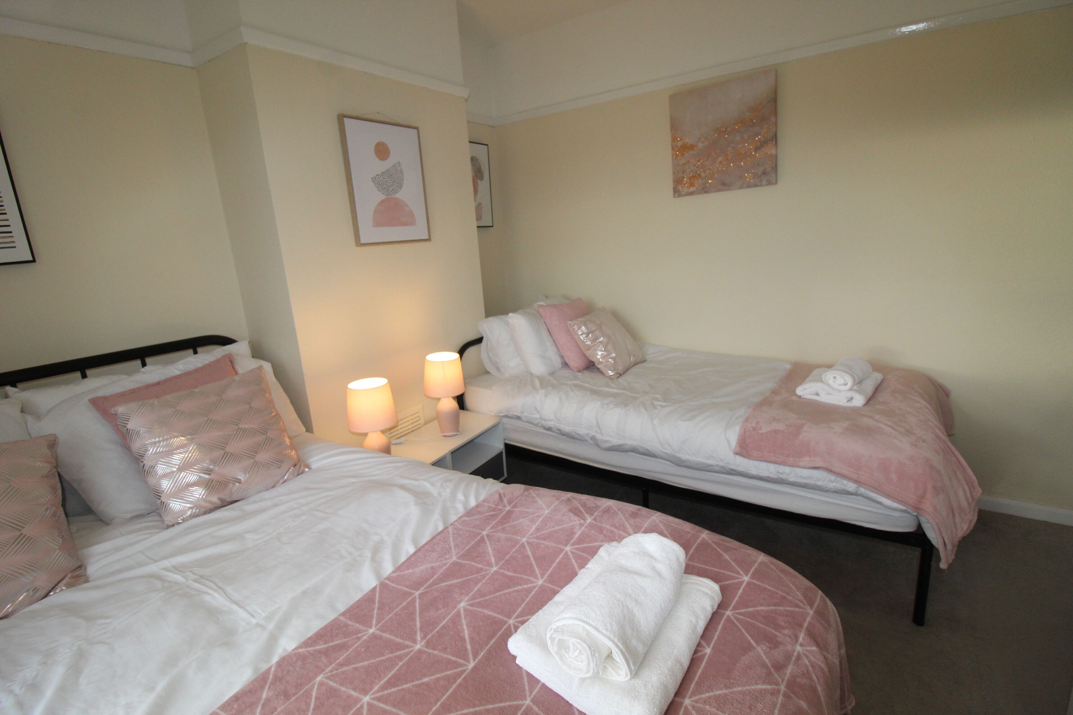 Una camera d'albergo con due letti, entrambi con biancheria rosa e bianca, un comodino con una lampada e opere d'arte alle pareti.