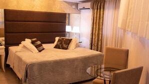 A hotel room with a large bed, a glass table, a chair, and a nightstand with a lamp. - Hotel Imigrantes (Bento Gonçalves)
