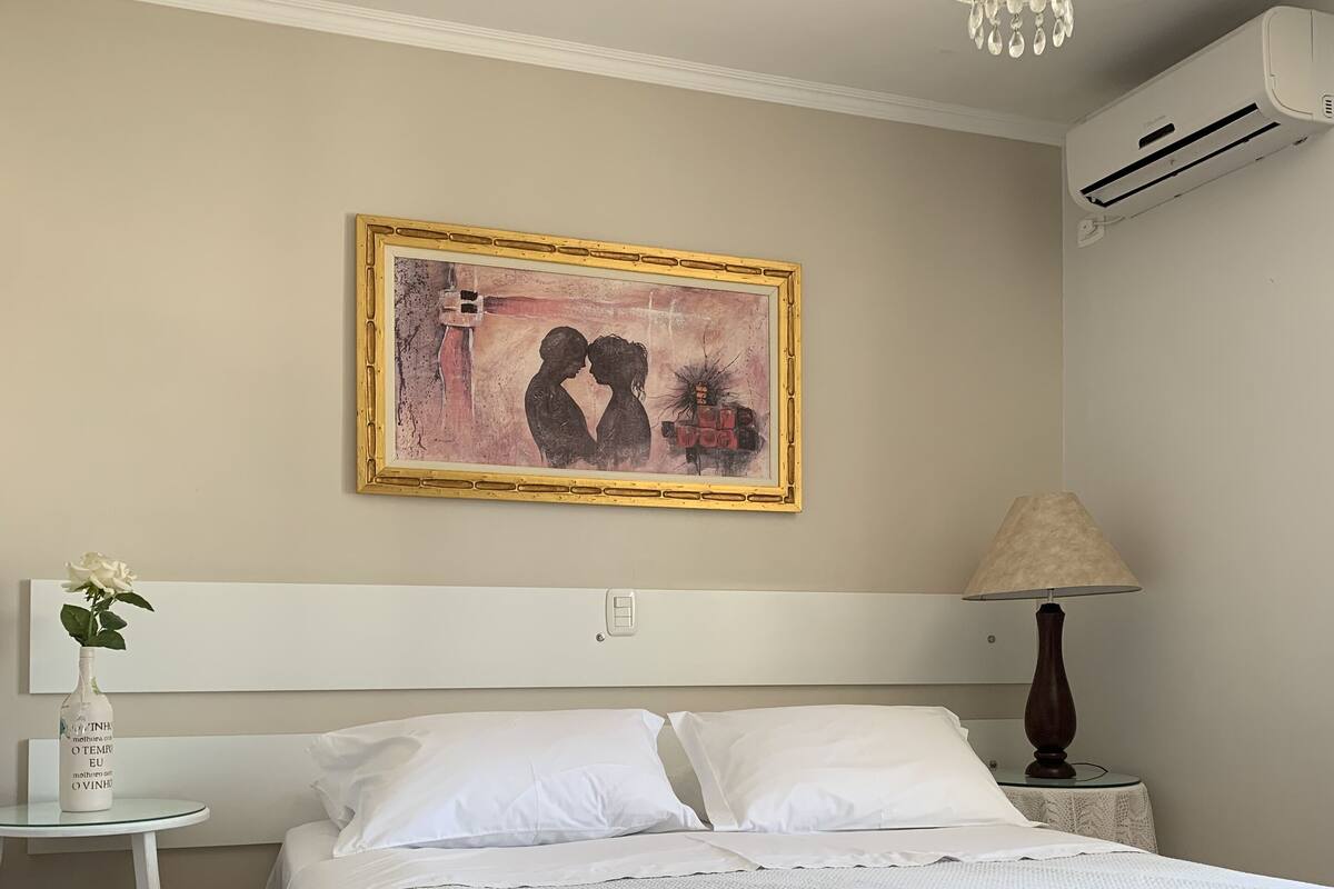 A neatly made bed with white linens, a bedside table with a vase of flowers, a wall-mounted air conditioner, and a framed artwork above the bed.