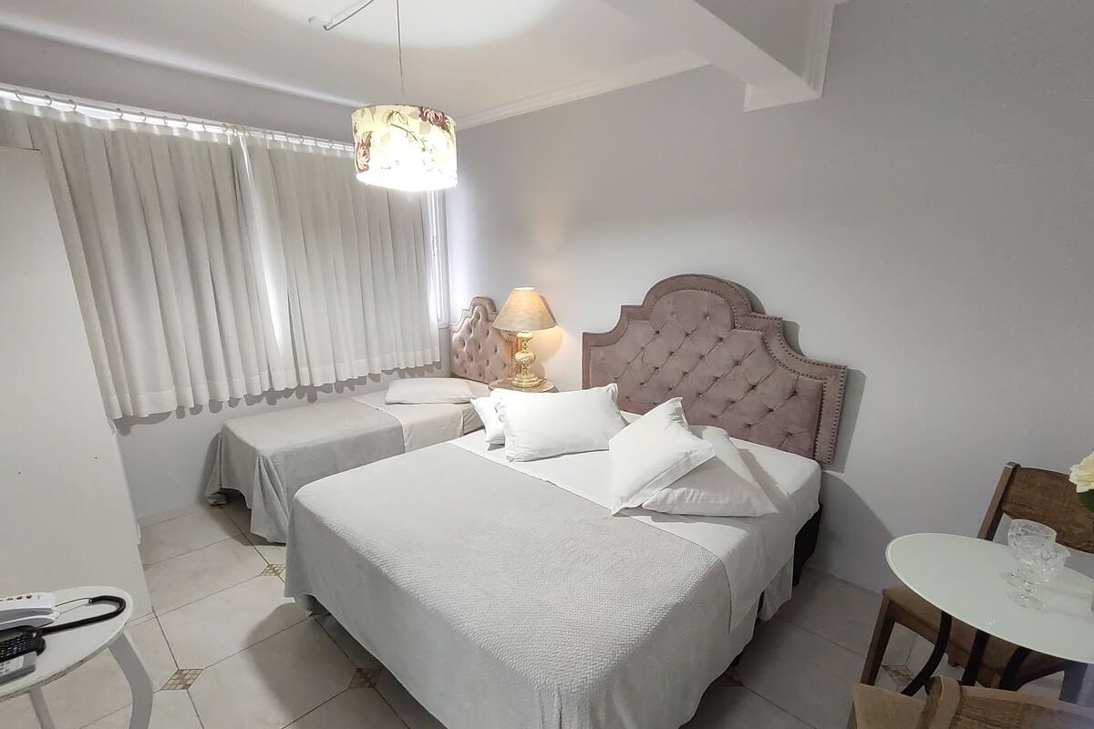 A hotel room with two beds, a bedside table with a lamp, a small round table with a chair, and a window with curtains.
