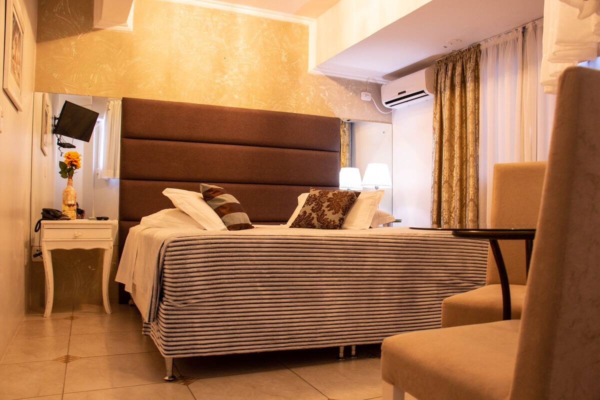 A hotel room with a large bed, a bedside table, a chair, a TV mounted on the wall, and a window with curtains.