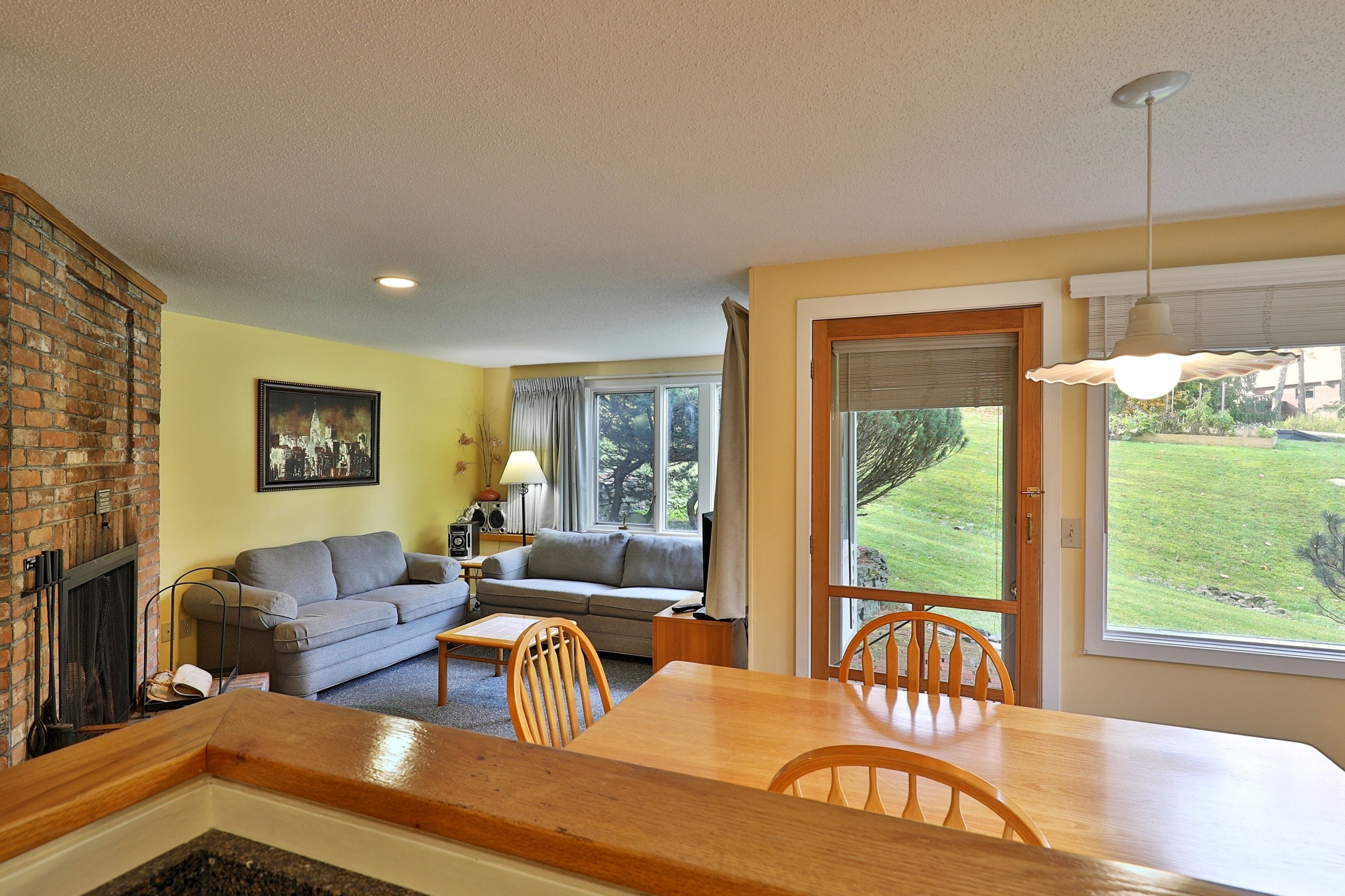 A living room with a fireplace, a sofa, a dining table with chairs, and a large window with a view of a grassy yard.