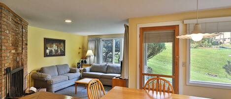 A living room with a fireplace, a sofa, a dining table with chairs, and a large window with a view of a grassy yard.