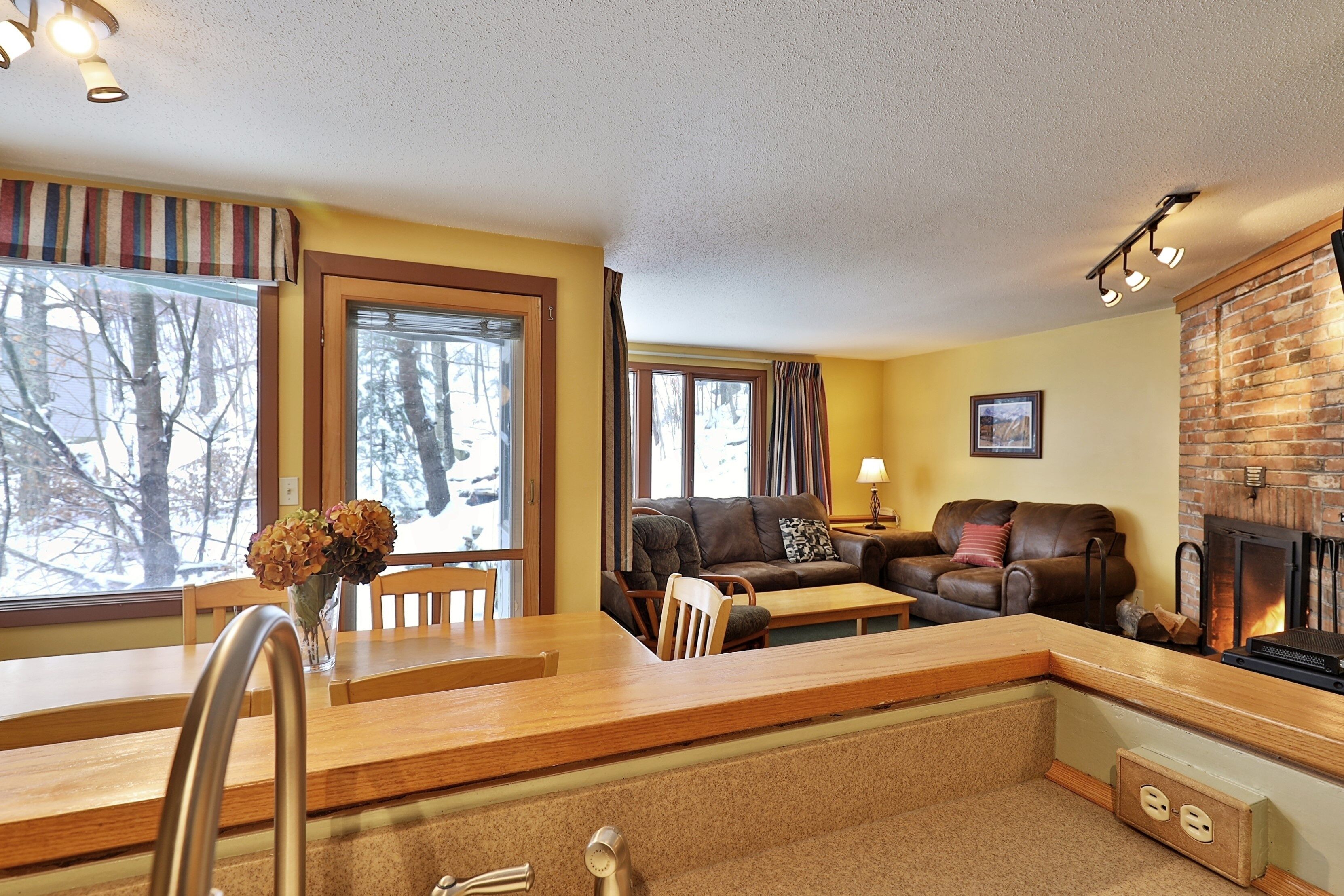 A cozy living room with a fireplace, wooden furniture, and large windows offering a view of trees.