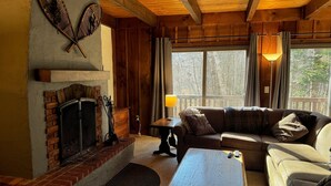 A cozy living room with a fireplace, wooden beams, a sofa, and a view through large windows. - Hemlock Ridge 2b: Charming Two Floor Condo In The Heart Of Killington. Perfect! 3 Bedroom Condo (Killington)
