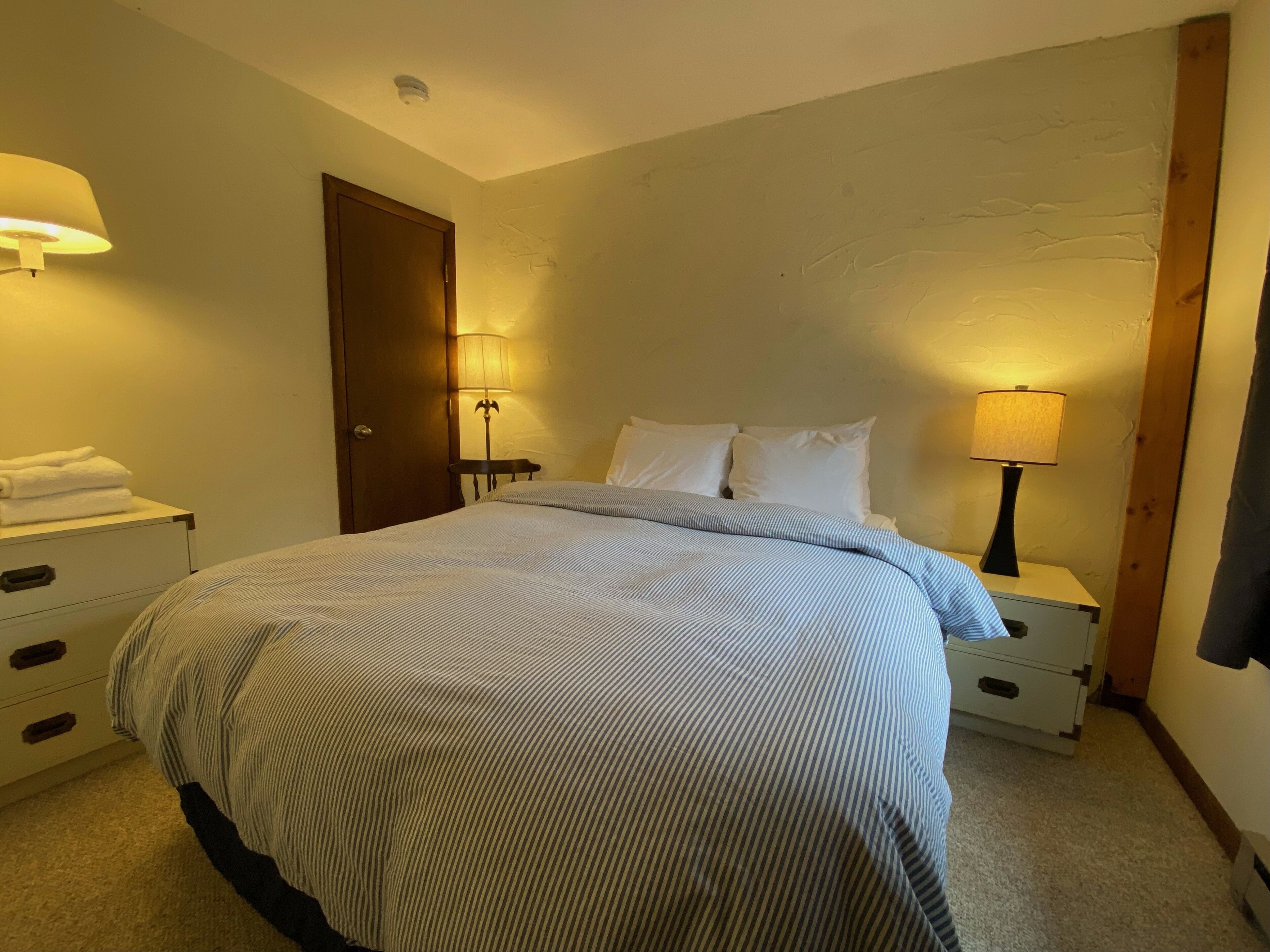 A neatly made bed with striped bedding, two bedside tables with lamps, a wooden door, and a wall with a textured finish.