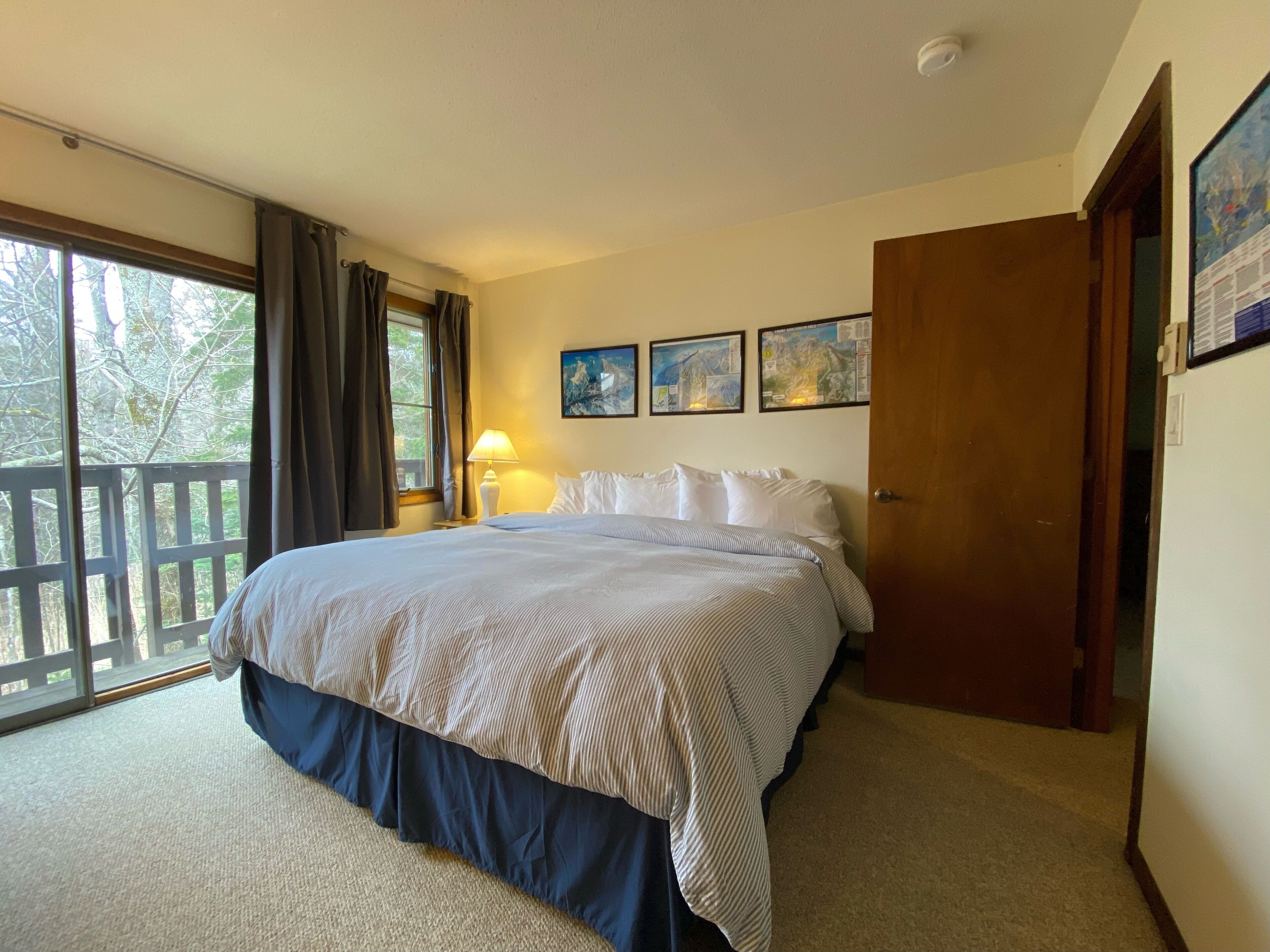 A bedroom with a large bed, a window with curtains, a nightstand with a lamp, and a door.
