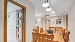 Un espace repas avec une table et des chaises en bois, un luminaire moderne et une vue sur la pièce adjacente, accessible par une porte ouverte.