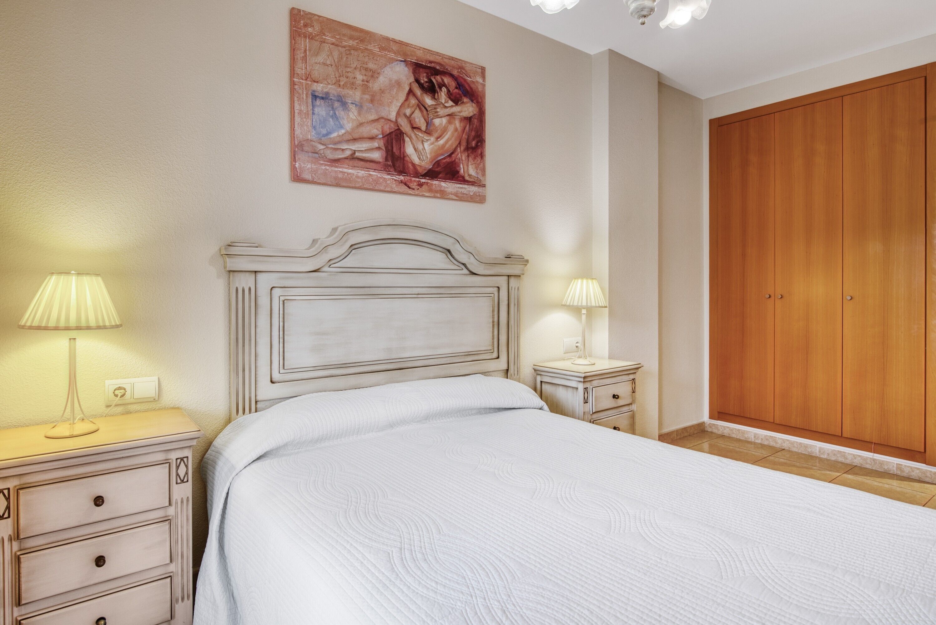 A bedroom with a large bed, two bedside tables with lamps, a wooden wardrobe, and a painting on the wall.