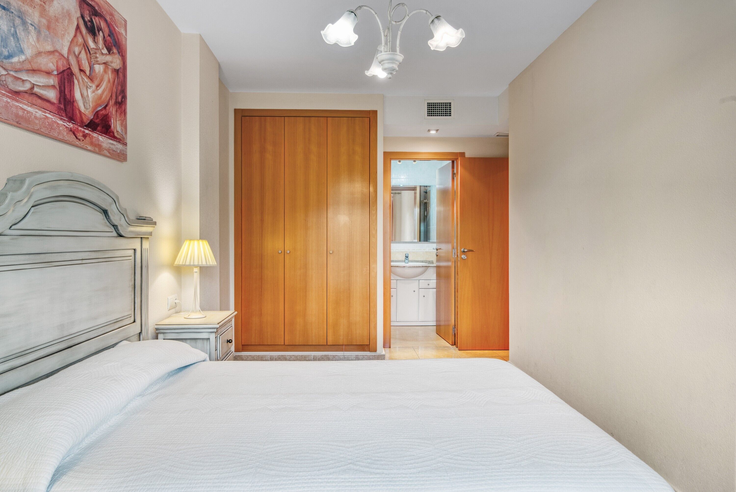 A bedroom with a bed, a nightstand, a lamp, a wooden wardrobe, and a bathroom visible through an open door.