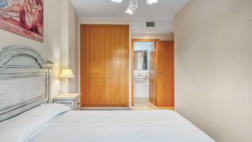 A bedroom with a bed, a nightstand, a lamp, a wooden wardrobe, and a bathroom visible through an open door.