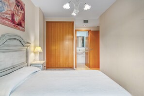Quarto com cama, mesa de cabeceira, abajur, guarda-roupa de madeira e banheiro visível por uma porta aberta.