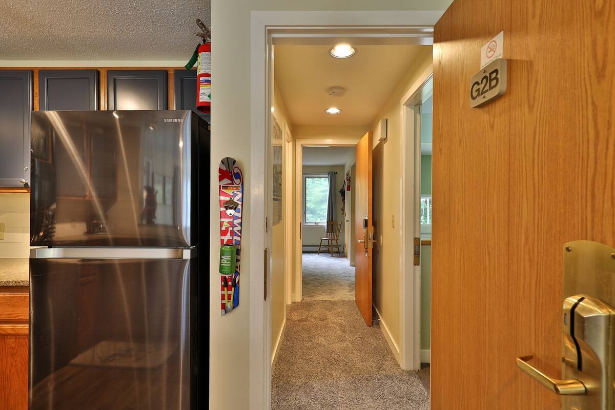 A modern kitchen with a stainless steel refrigerator, a wooden door with a G2B sign, and a hallway leading to a living area.