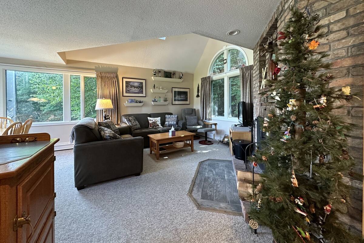 A cozy living room with a fireplace, a Christmas tree, and a large window offering a view of the outdoors.