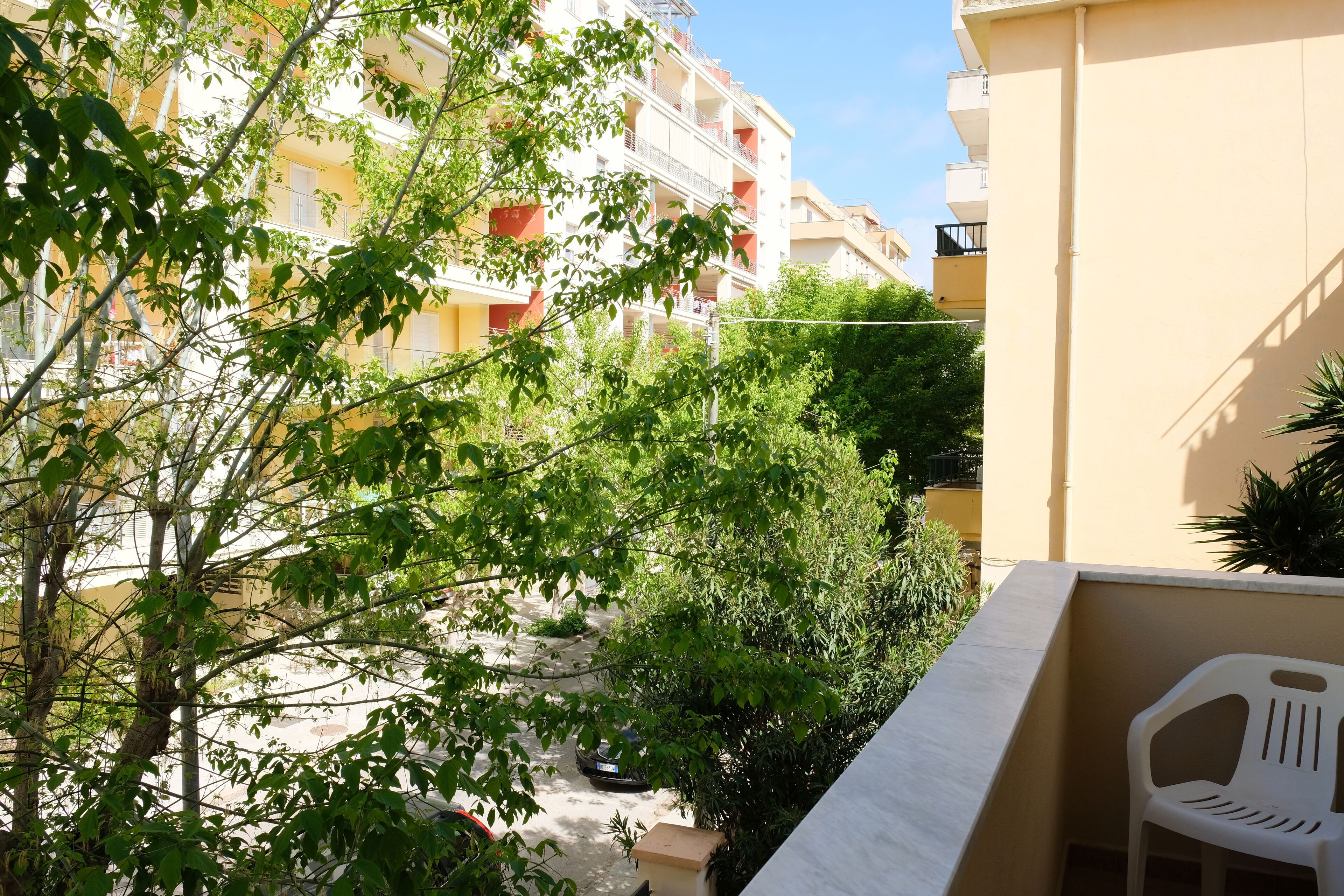 Ein Balkon mit einem weißen Plastikstuhl, der auf einen Innenhof mit Bäumen und einem Gebäude im Hintergrund blickt.