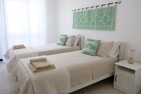 A hotel room with two beds, each with a beige bedspread and pillows, a small bedside table with a lamp, and a wall-mounted towel rack. - Just 80 meters from the sea, Best area of Alghero, 3 bedrooms+private parking+AC (Alghero)