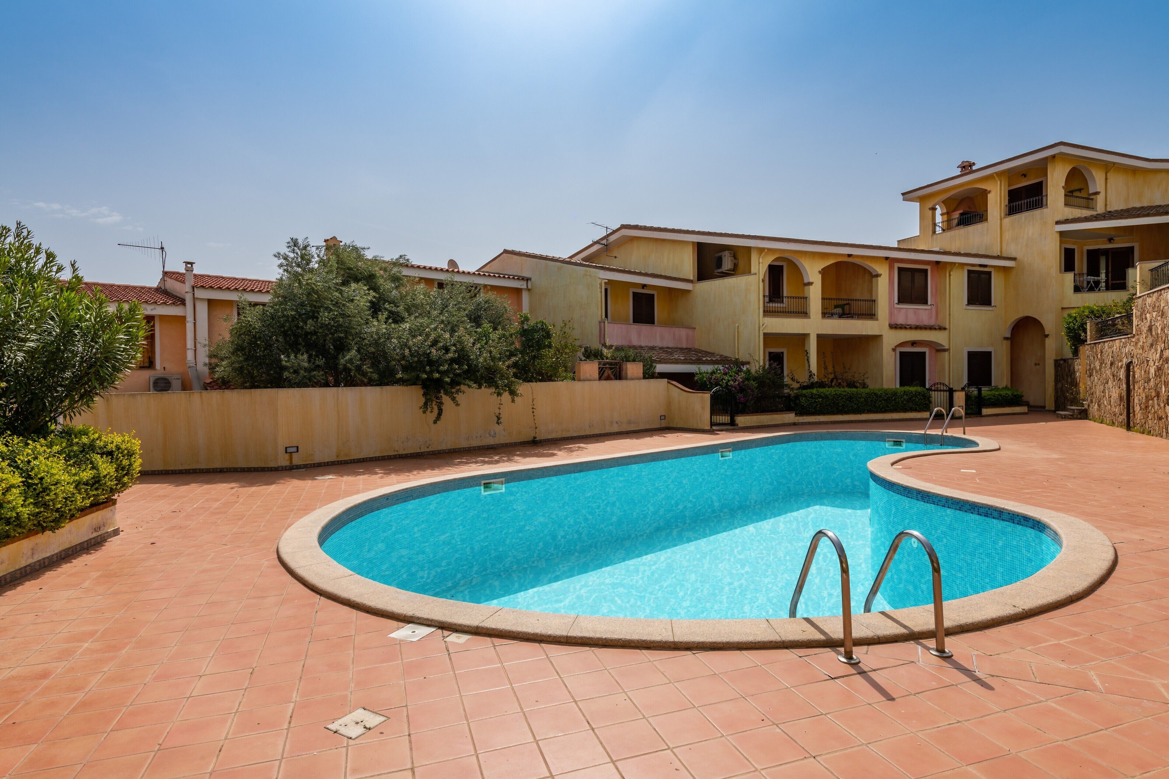 A swimming pool with a tiled deck, surrounded by multi-story buildings with balconies and a clear sky.
