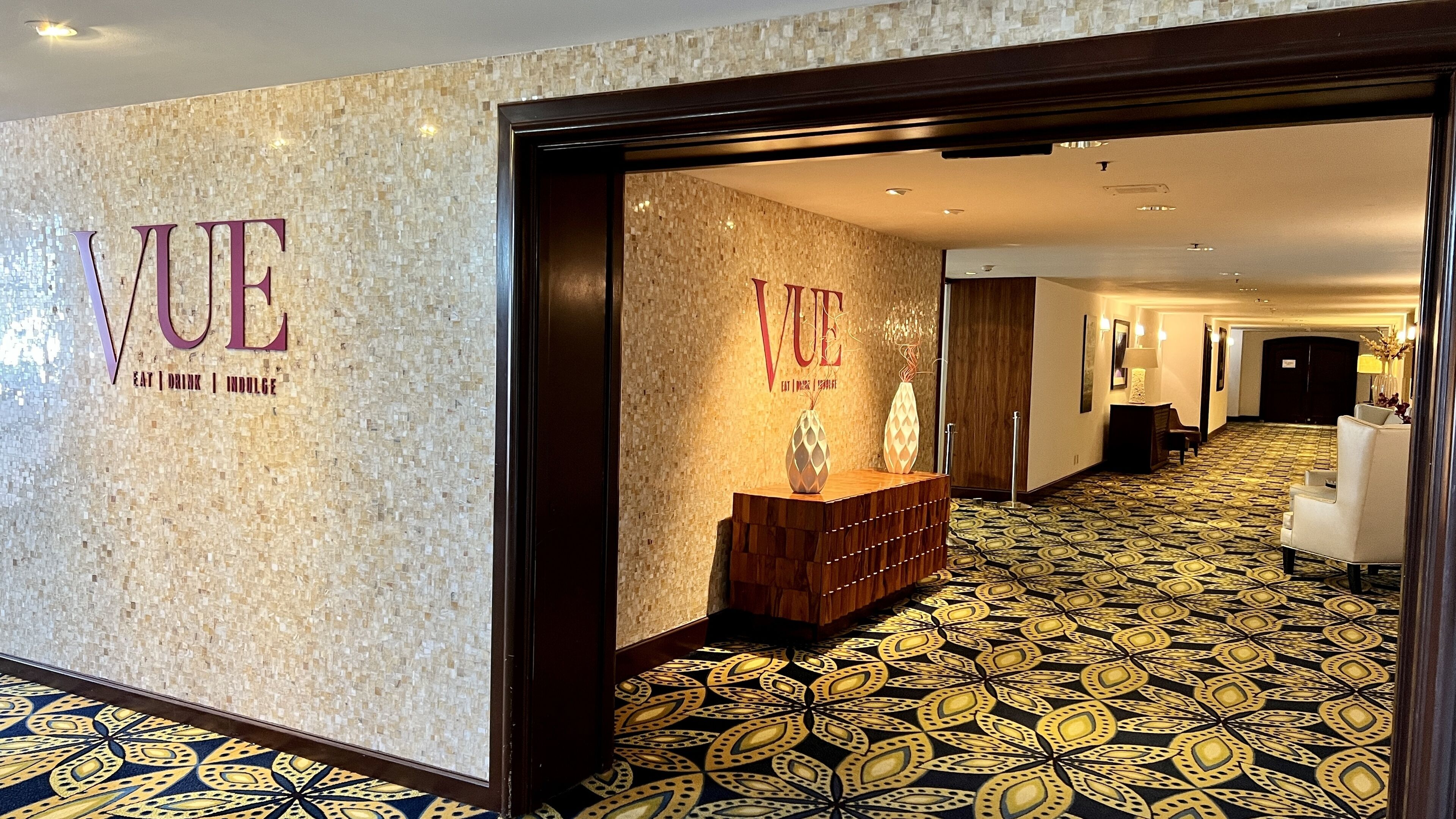 Ein Hotelkorridor mit gemustertem Teppich, einem Empfangstresen und 'VUE'-Beschilderung an den Wänden.