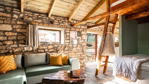 Una habitación rústica con paredes de piedra, techo de madera, un sofá verde con cojines, una mesa de centro de madera y una cama con una manta blanca.