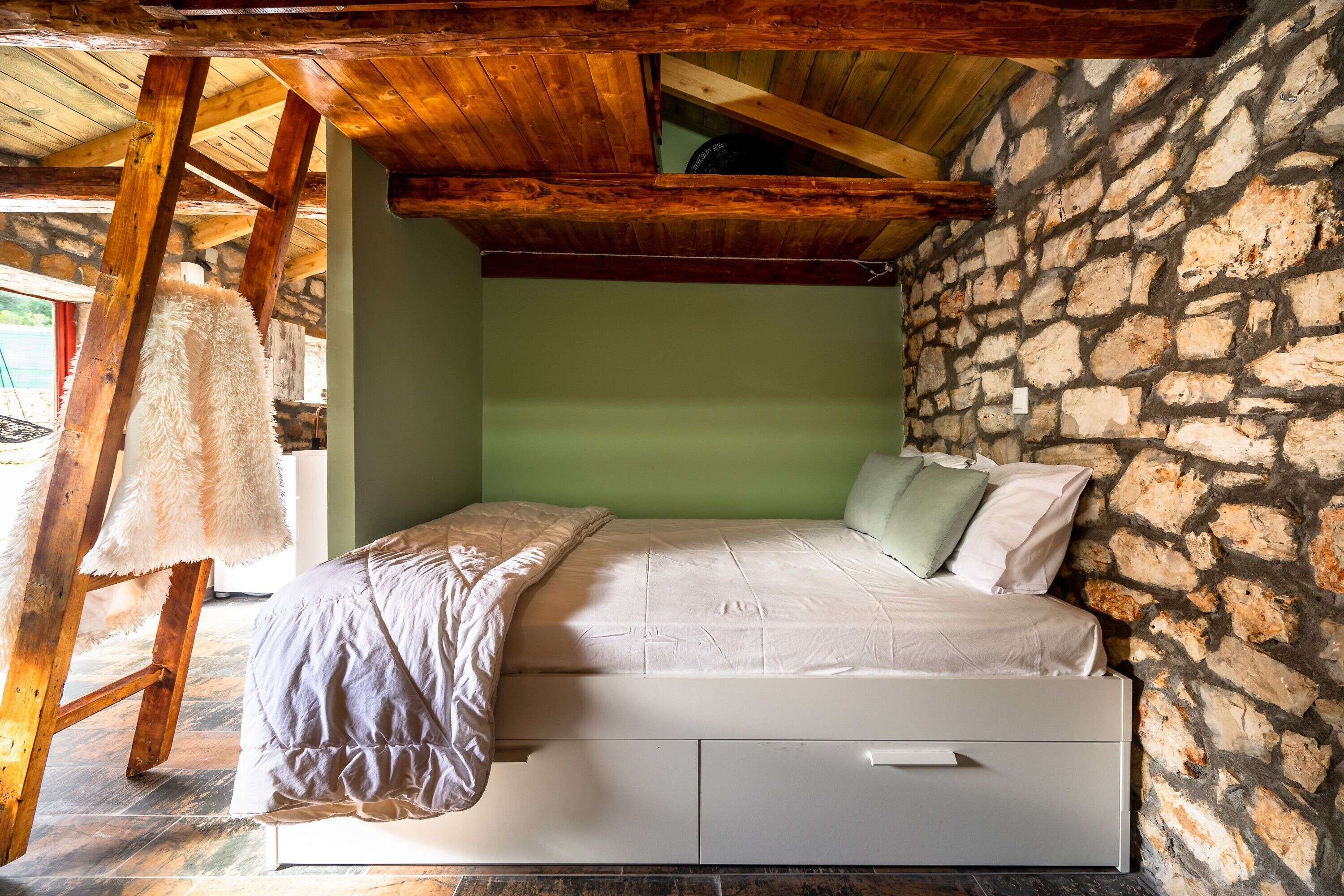 Un dormitorio con pared de piedra, vigas de madera en el techo, una cama con colchón y almohadas blancas, y una silla de madera.
