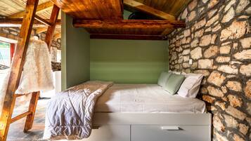 Un dormitorio con pared de piedra, vigas de madera en el techo, una cama con colchón y almohadas blancas, y una silla de madera.