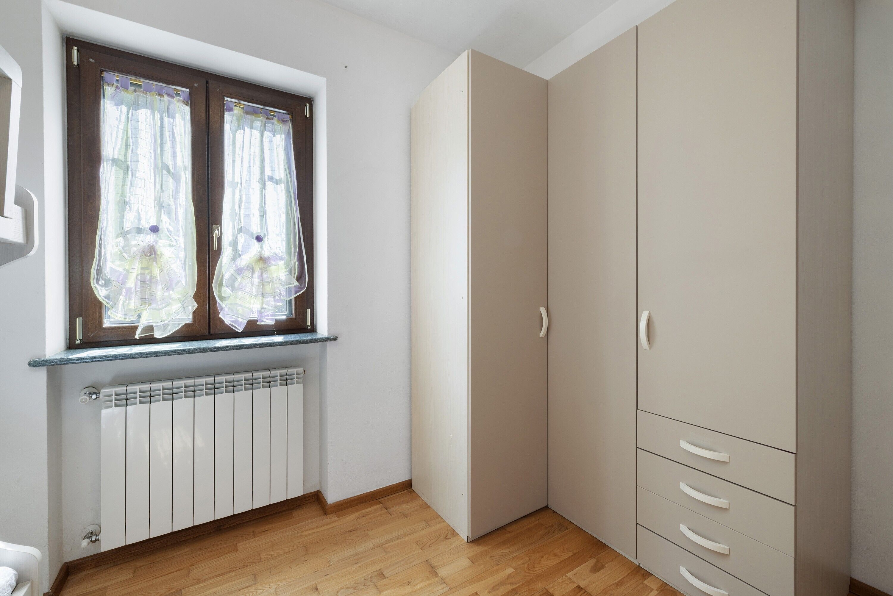 Ein Zimmer mit Fenster, Heizkörper, Holzboden und einem Schrank mit Schubladen.