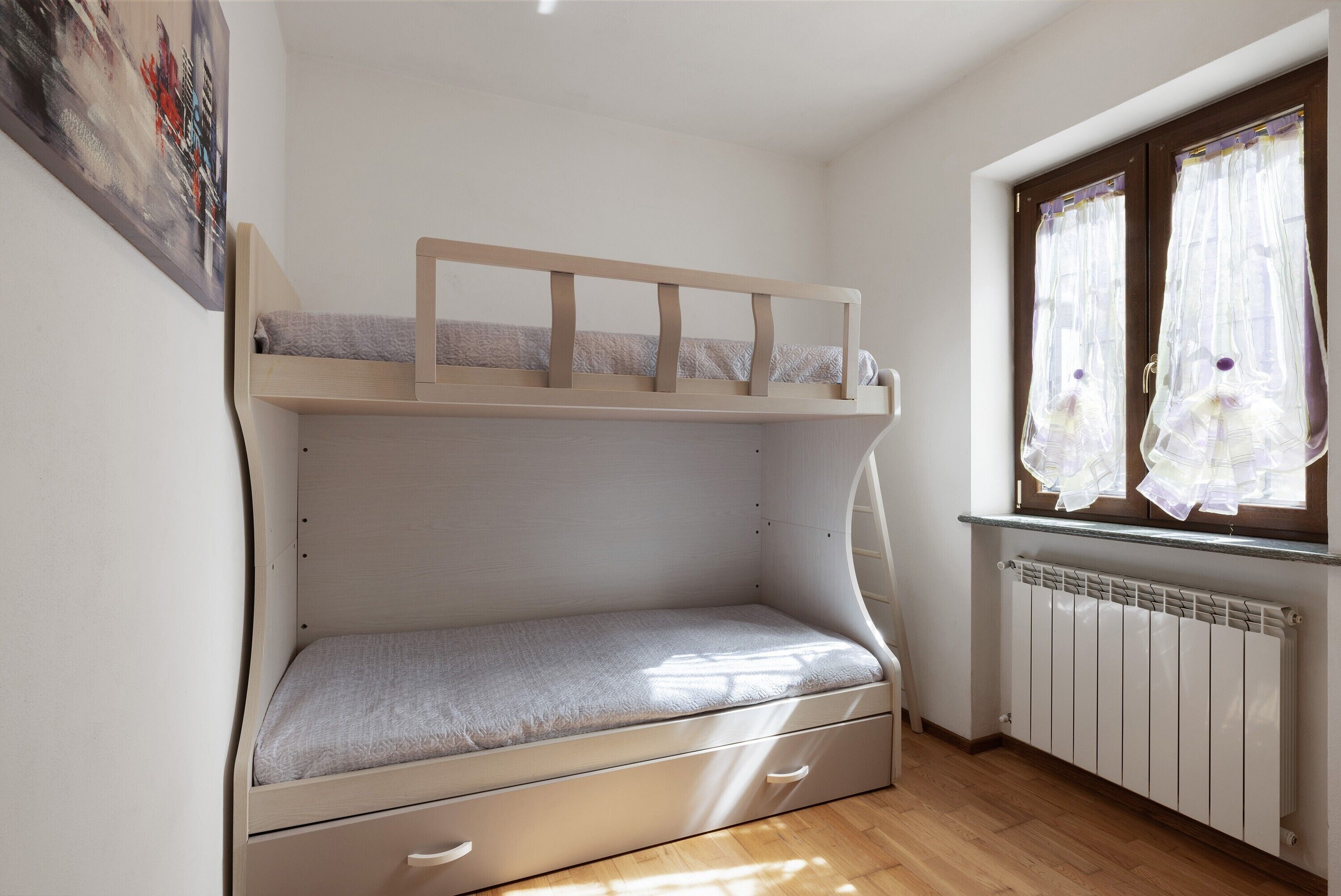 Een stapelbed met een onderbed, een lade, een radiator en een raam met gordijnen.