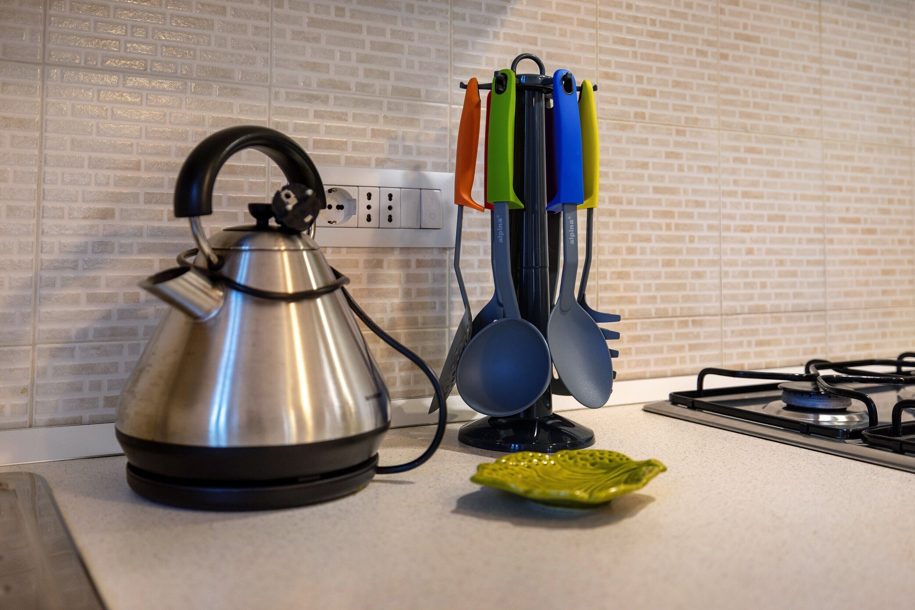 A kitchen with a kettle, a set of colorful ladles, and a green plate on the counter.