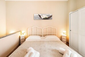A neatly made bed with a heart-patterned blanket, a wall-mounted picture of flowers in a boat, and two bedside tables with lamps.