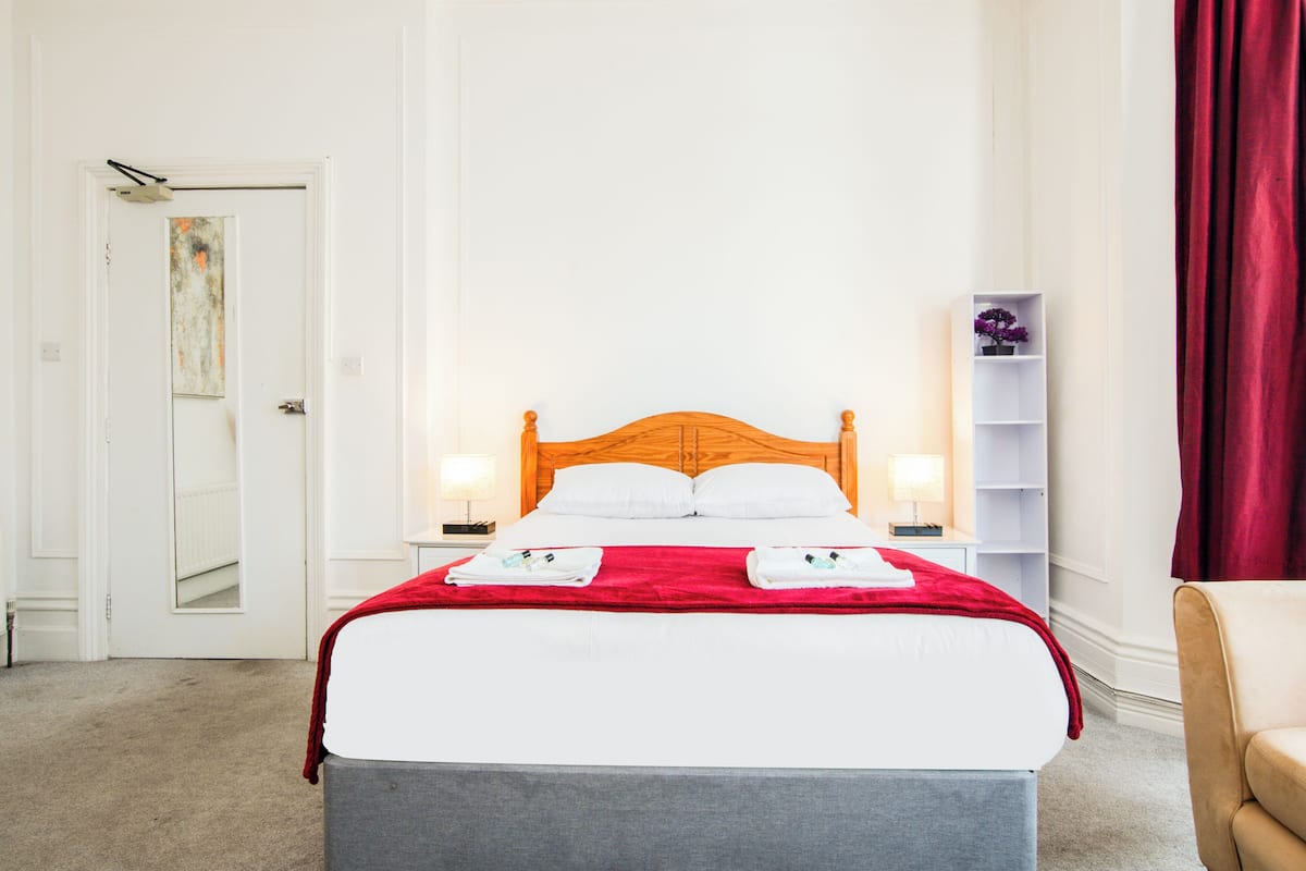 Una habitación de hotel con una cama, dos lámparas de noche, una manta roja, un banco gris, un estante blanco con una flor morada y una cortina roja.