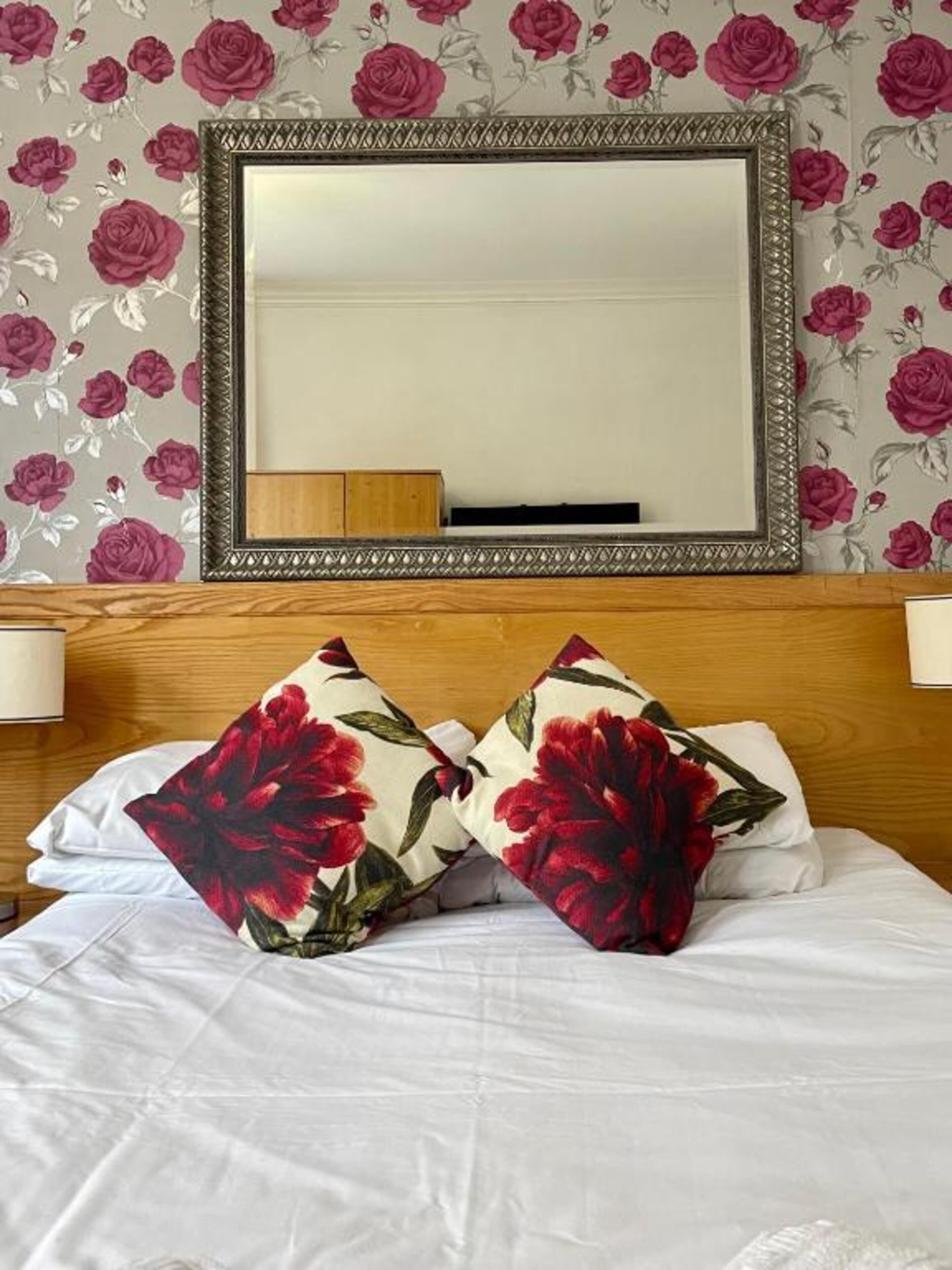 A hotel room with a bed, two pillows with a red floral pattern, a large mirror above the bed, and floral wallpaper on the walls.