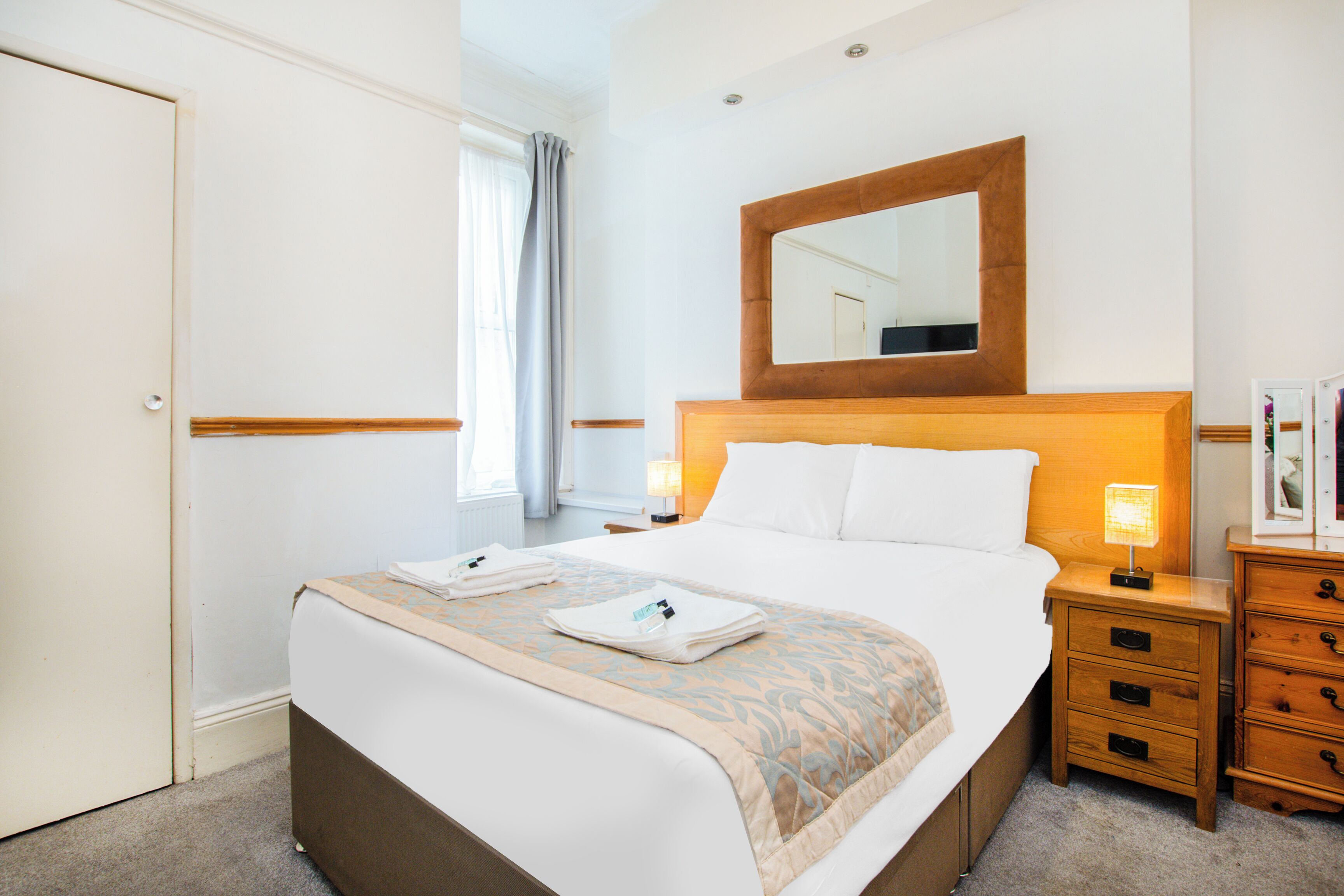A neatly made bed with a wooden headboard, a bedside table with a lamp, and a mirror above the bed.