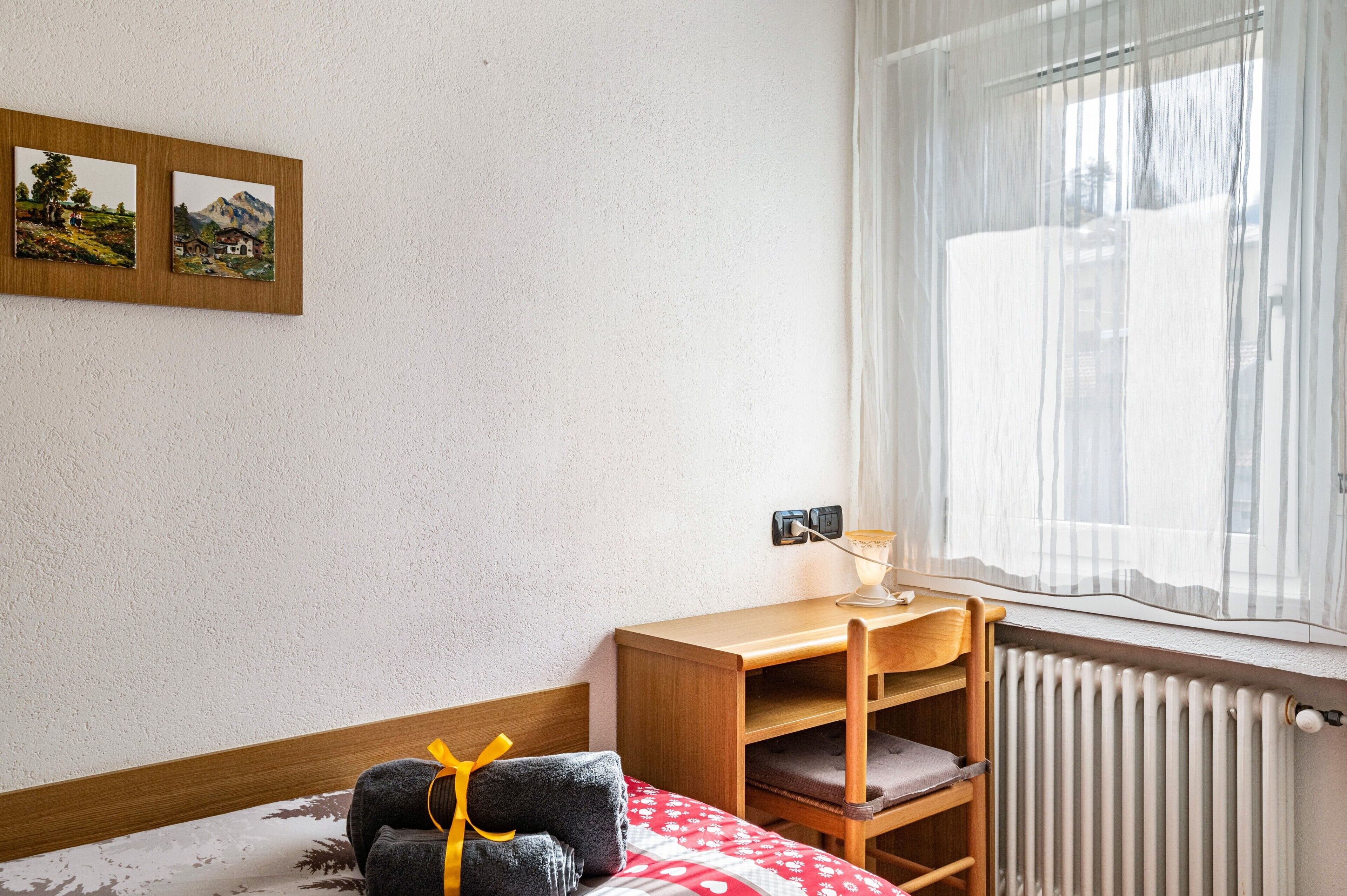 A hotel room with a bed, a wooden desk with a lamp, a radiator, and two framed pictures on the wall.